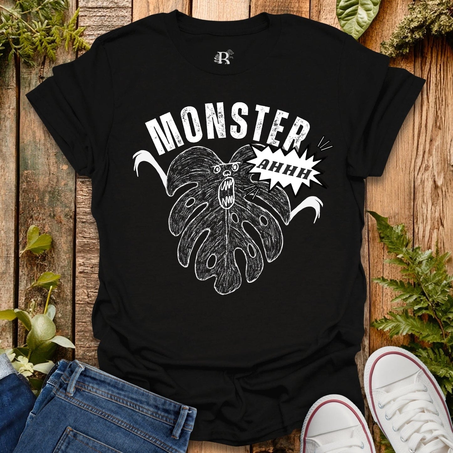 Black t-shirt with a graphic design of a monster and 'MONSTER AHHH' text on a wooden surface with plants.