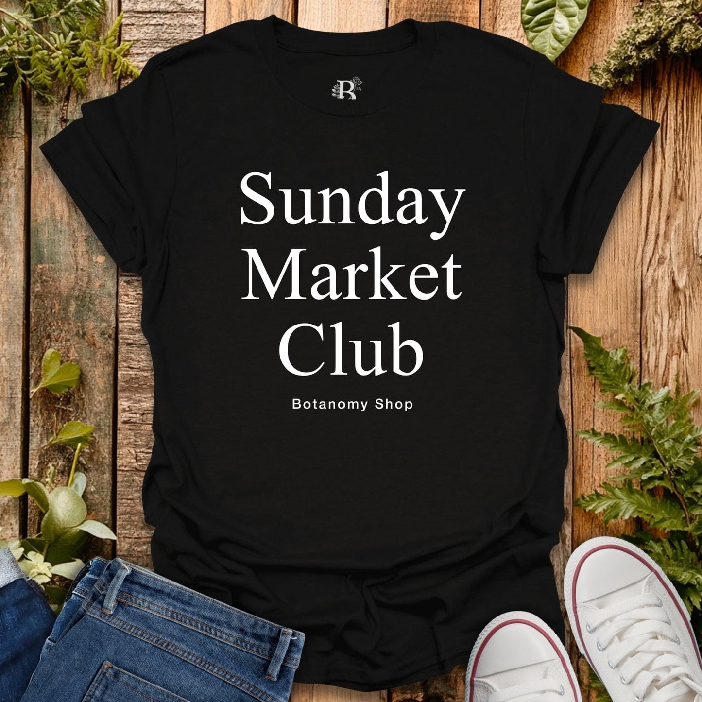 Black t-shirt with 'Sunday Market Club' text on a wooden surface with plants.