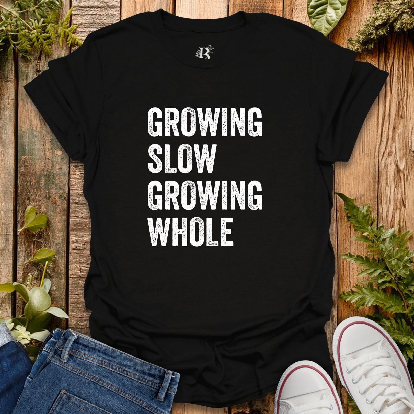 Black t-shirt with 'GROWING SLOW GROWING WHOLE' text on a wooden surface with plants.