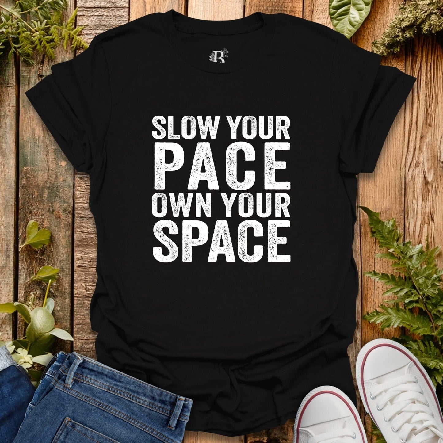 Black t-shirt with 'Slow your pace, own your space' text on a wooden surface with plants.