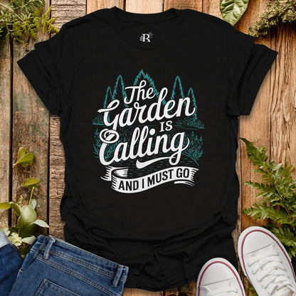 Black t-shirt with 'The Garden is Calling and I Must Go' text on a wooden surface with plants.