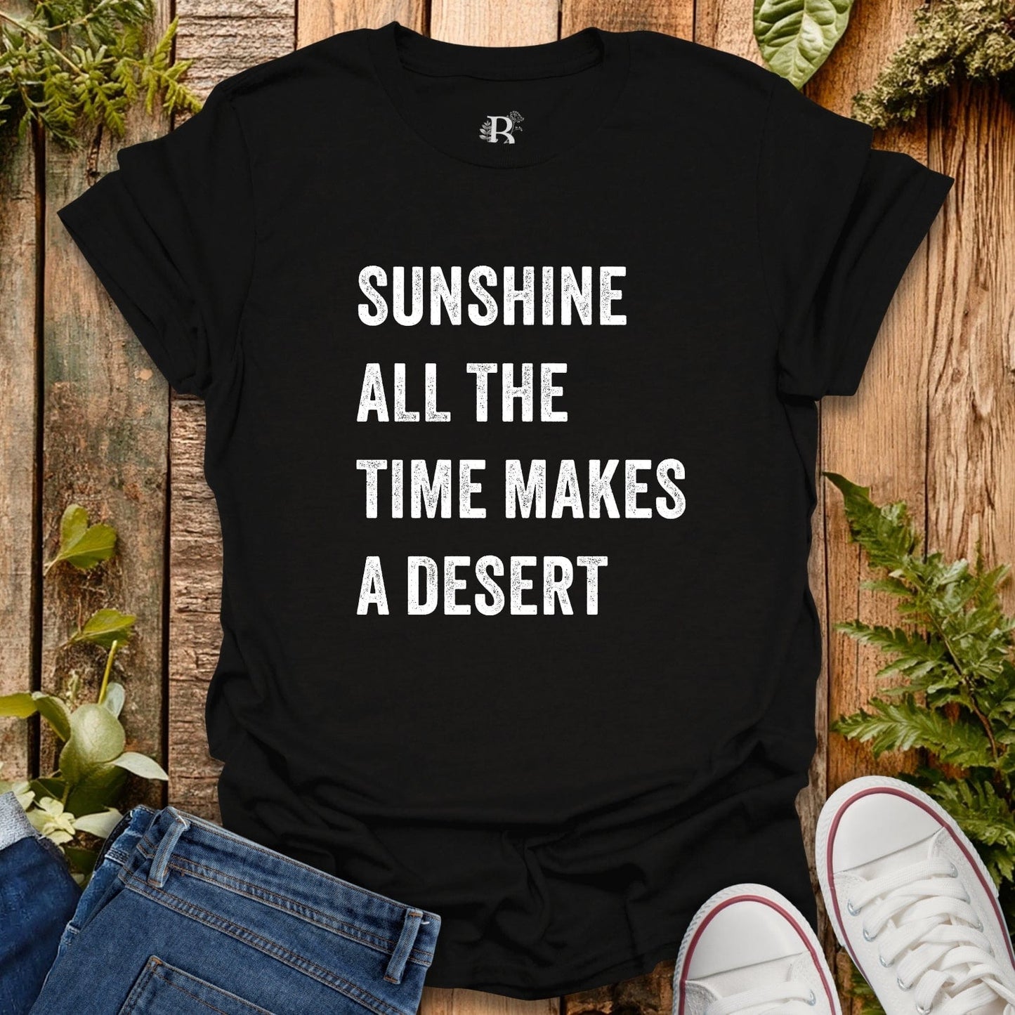 Black t-shirt with white text 'Sunshine all the time makes a desert' on a wooden surface with plants.