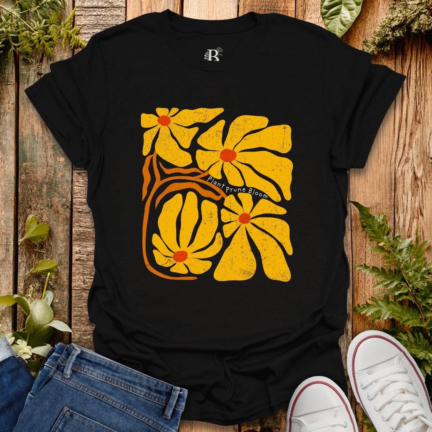 Black t-shirt with yellow floral design on a wooden surface with plants.