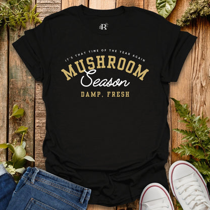 Black t-shirt with 'Mushroom Season' text on a wooden surface with plants.