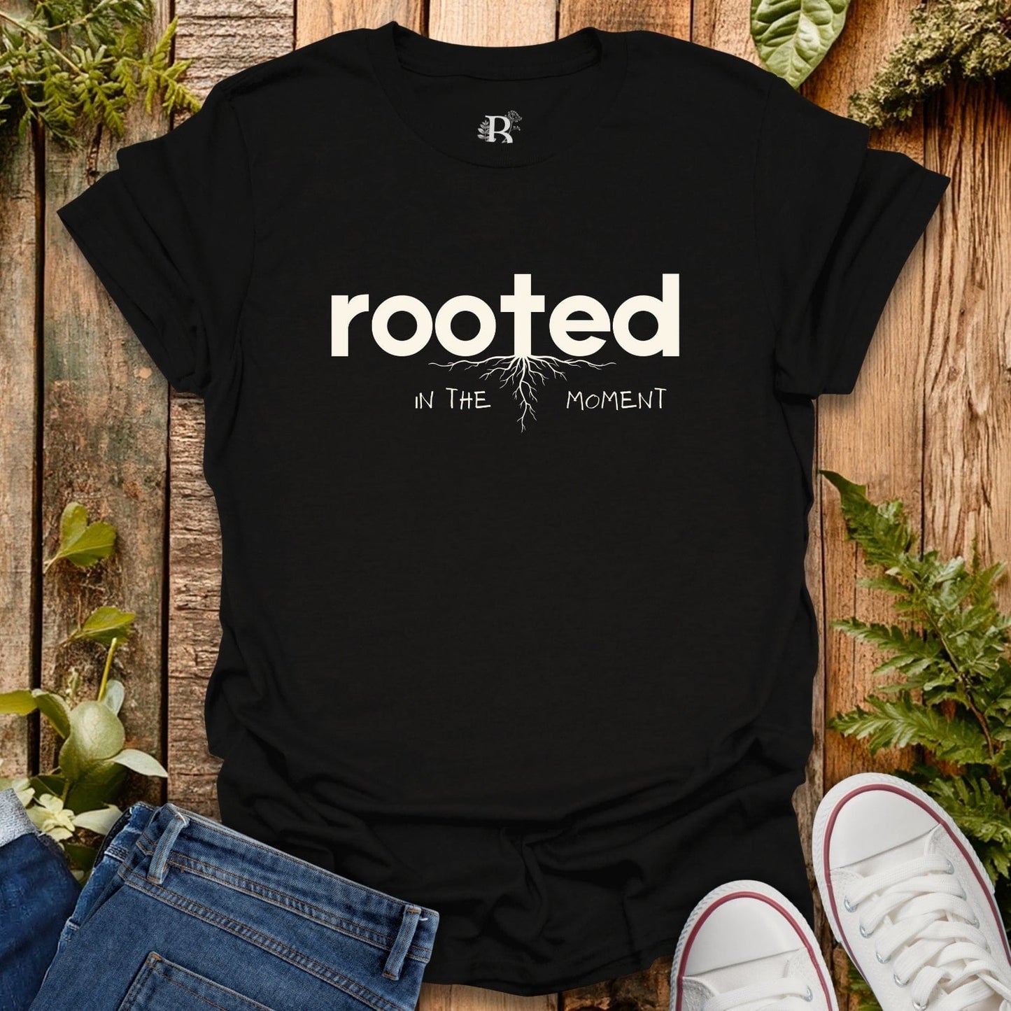 Black t-shirt with 'rooted in the moment' text on a wooden surface with plants.