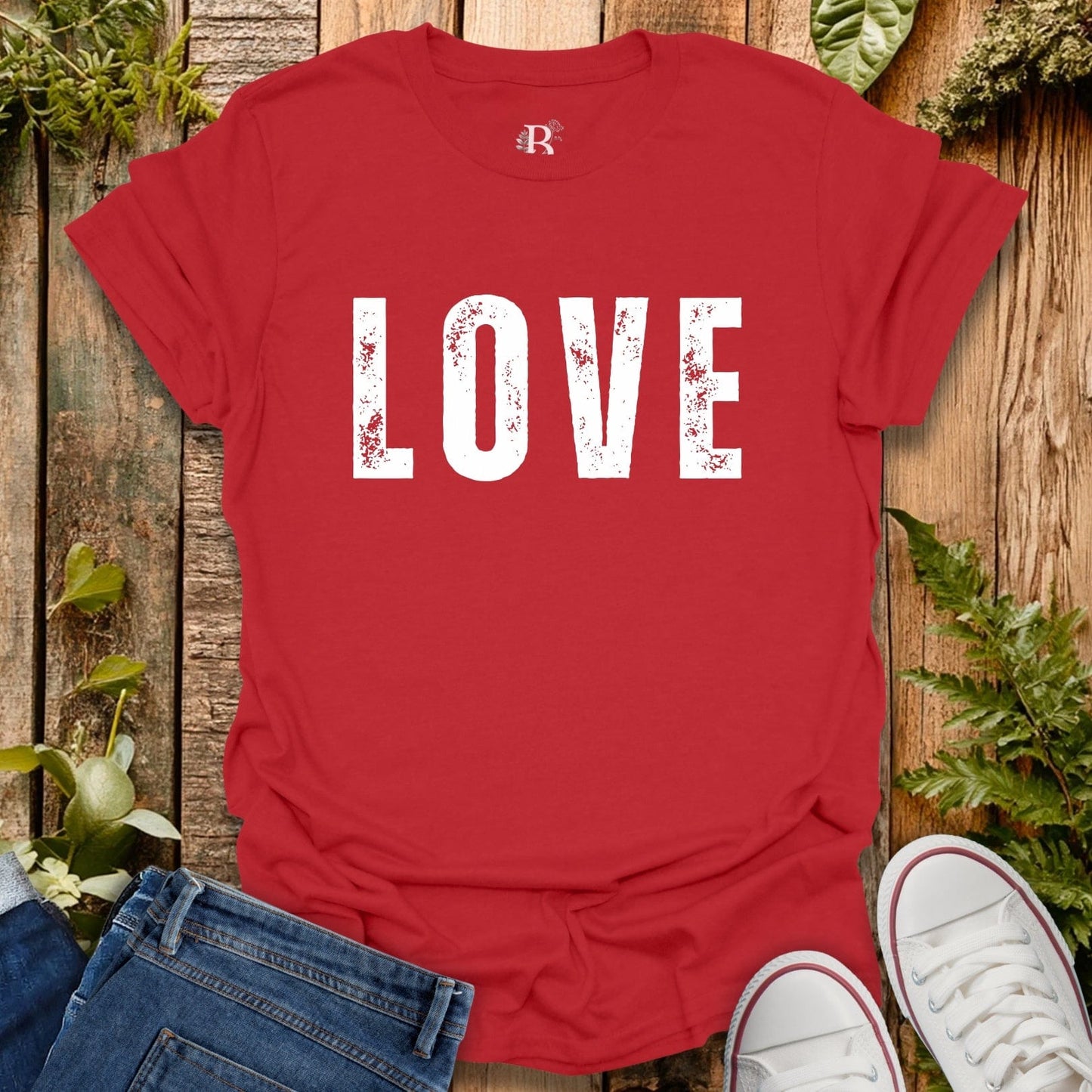 Red t-shirt with 'LOVE' printed on it, placed on a wooden surface with jeans and white sneakers.