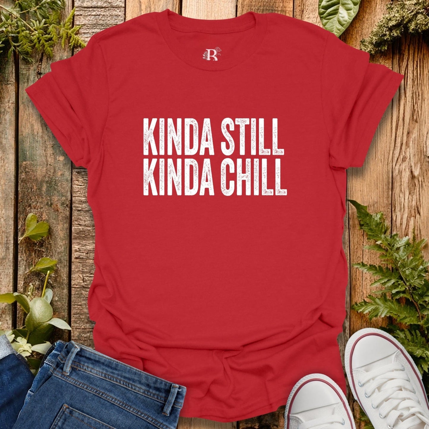 Red t-shirt with 'Kinda Still Kinda Chill' text on a wooden surface with jeans and sneakers.