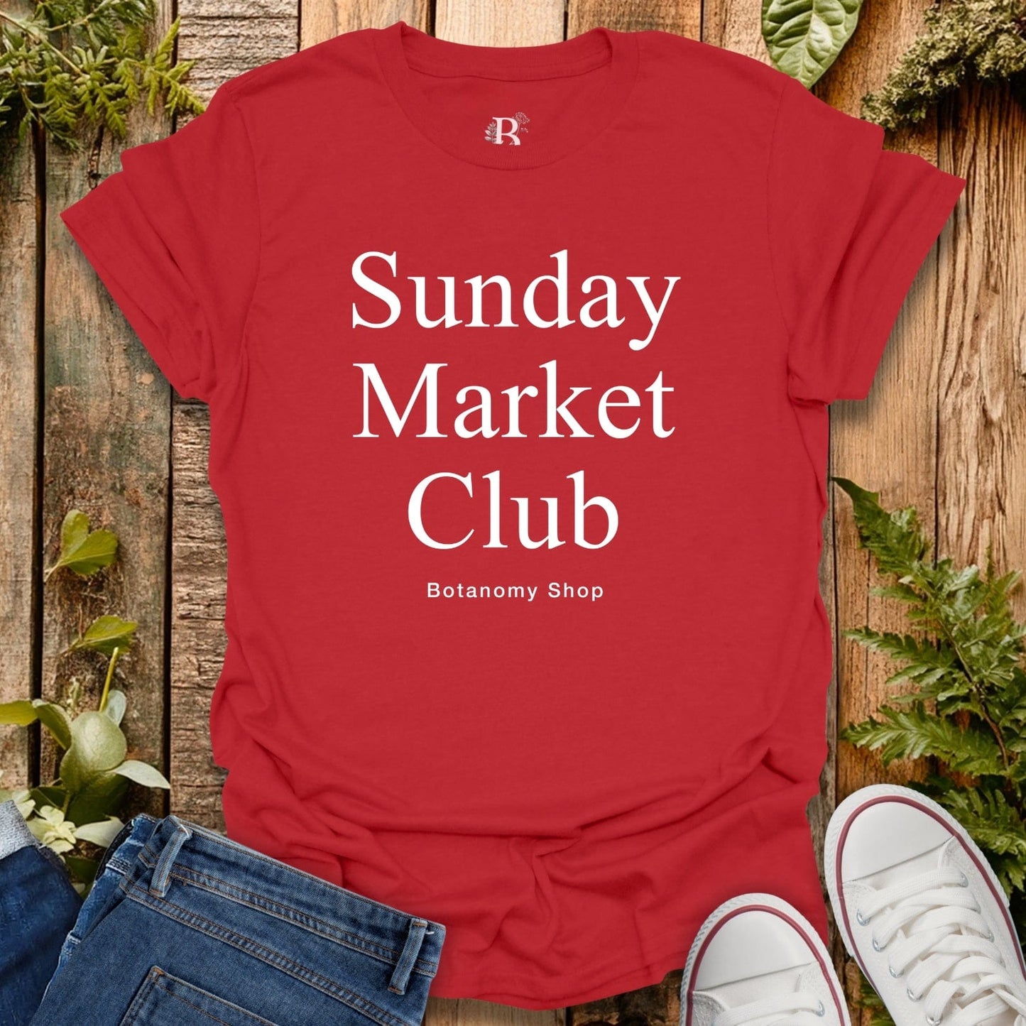 Red t-shirt with 'Sunday Market Club' text on a wooden surface with plants.