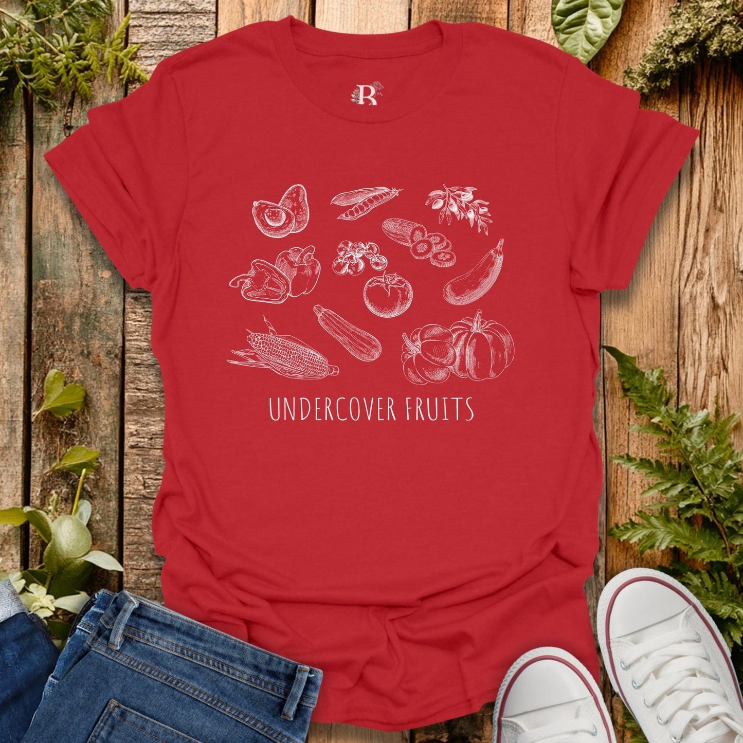 Red t-shirt with white graphic design of fruits and vegetables on a wooden surface with jeans and sneakers.