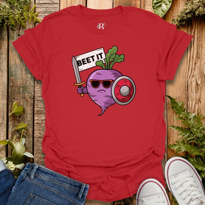Red t-shirt with a graphic of a beet holding a sword and shield, standing on a wooden surface with jeans and white sneakers.
