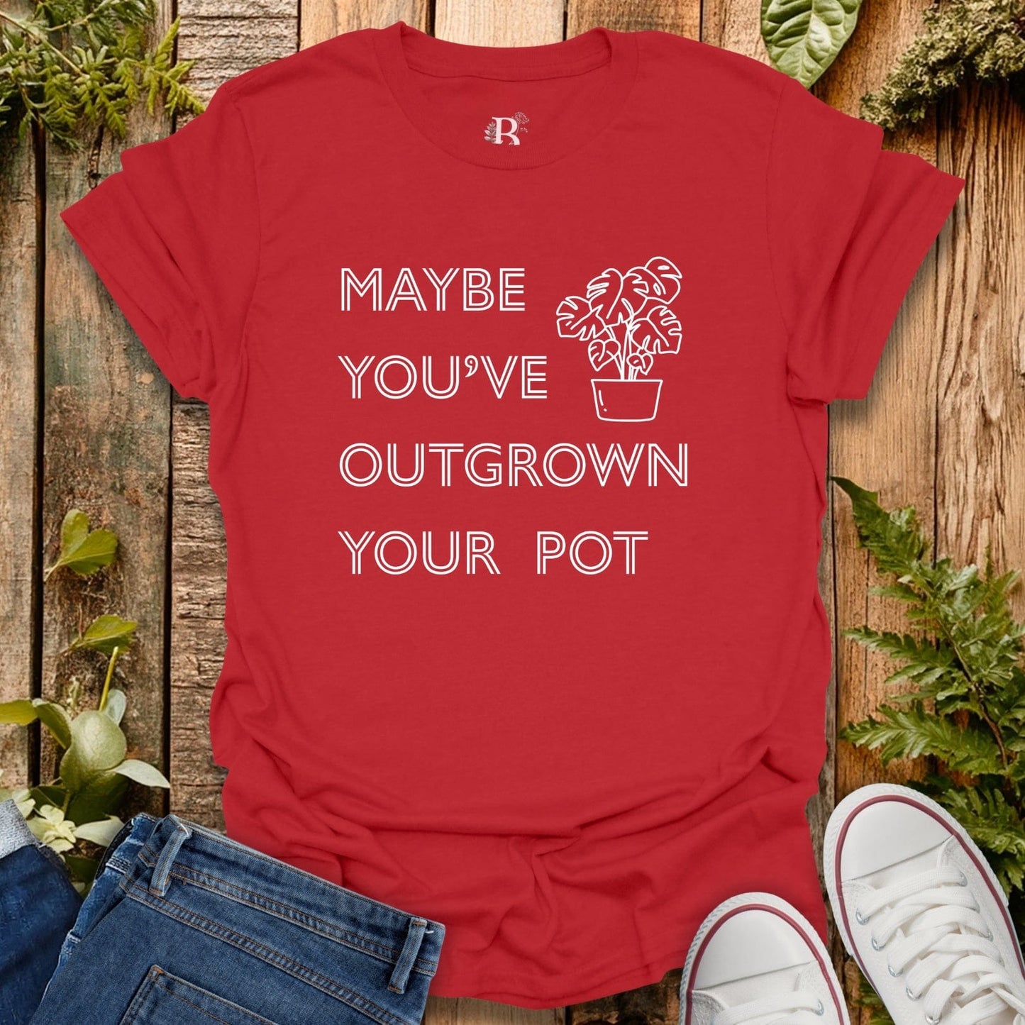 Red t-shirt with text and plant graphic on a wooden surface with plants and shoes.