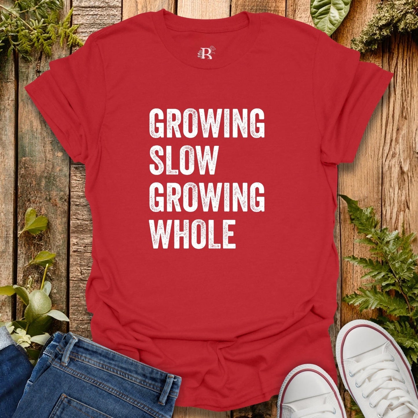 Red t-shirt with 'GROWING SLOW GROWING WHOLE' text on a wooden surface with jeans and sneakers.