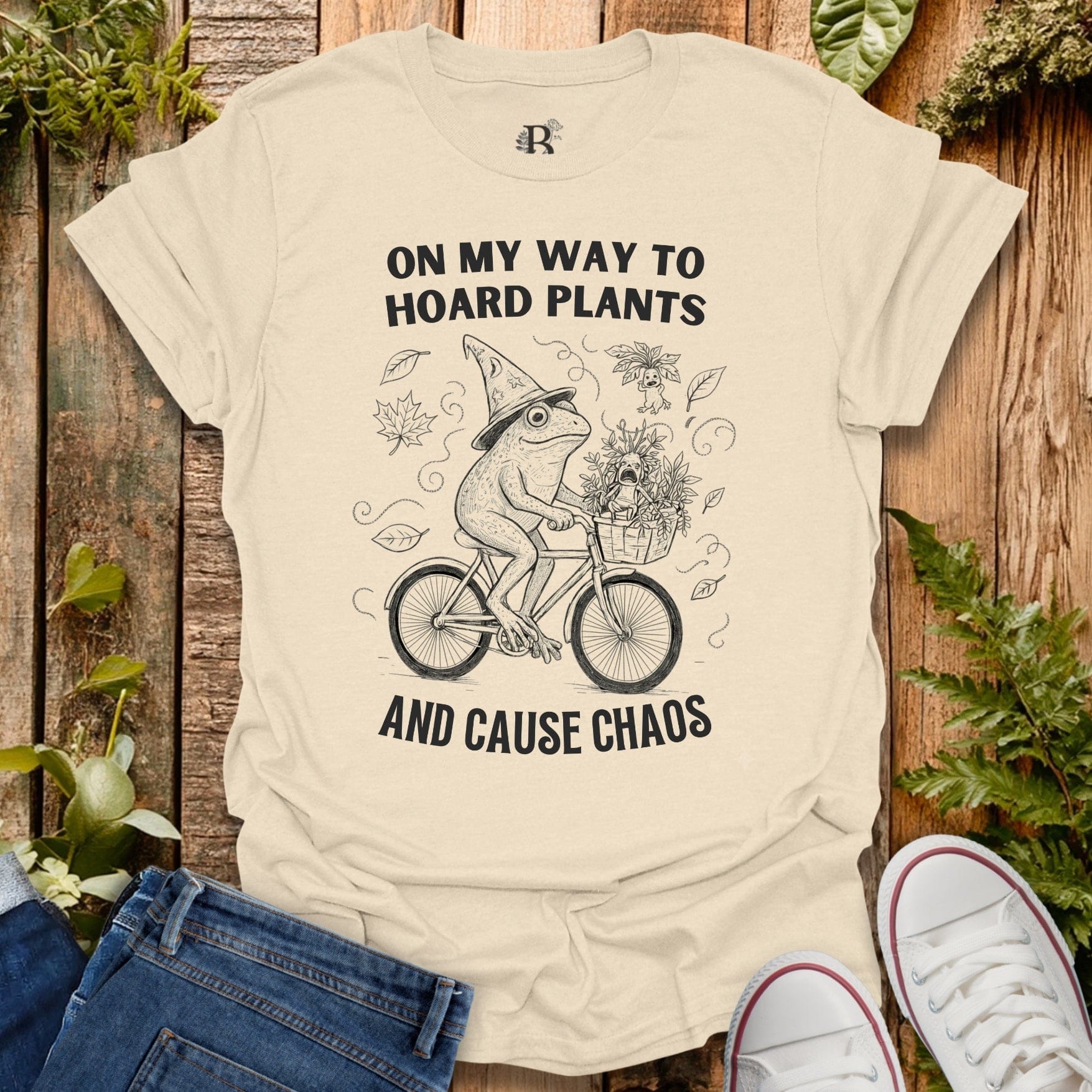 Beige t-shirt with a graphic of a frog riding a bike and text on a wooden background with plants.