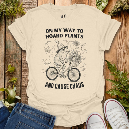 Beige t-shirt with a graphic of a frog riding a bike and text on a wooden background with plants.