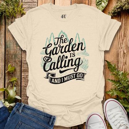 Beige t-shirt with 'The Garden is Calling and I Must Go' text on a wooden background with plants.