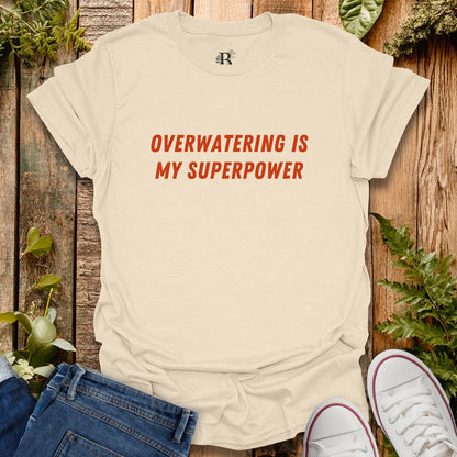 Beige t-shirt with 'Overwatering is my Superpower' text on a wooden background with plants.