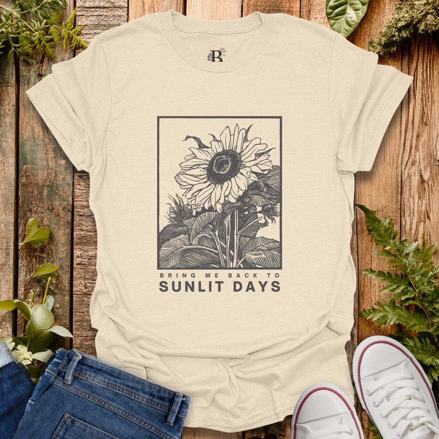 Beige t-shirt with sunflower graphic and text on a wooden surface with plants.