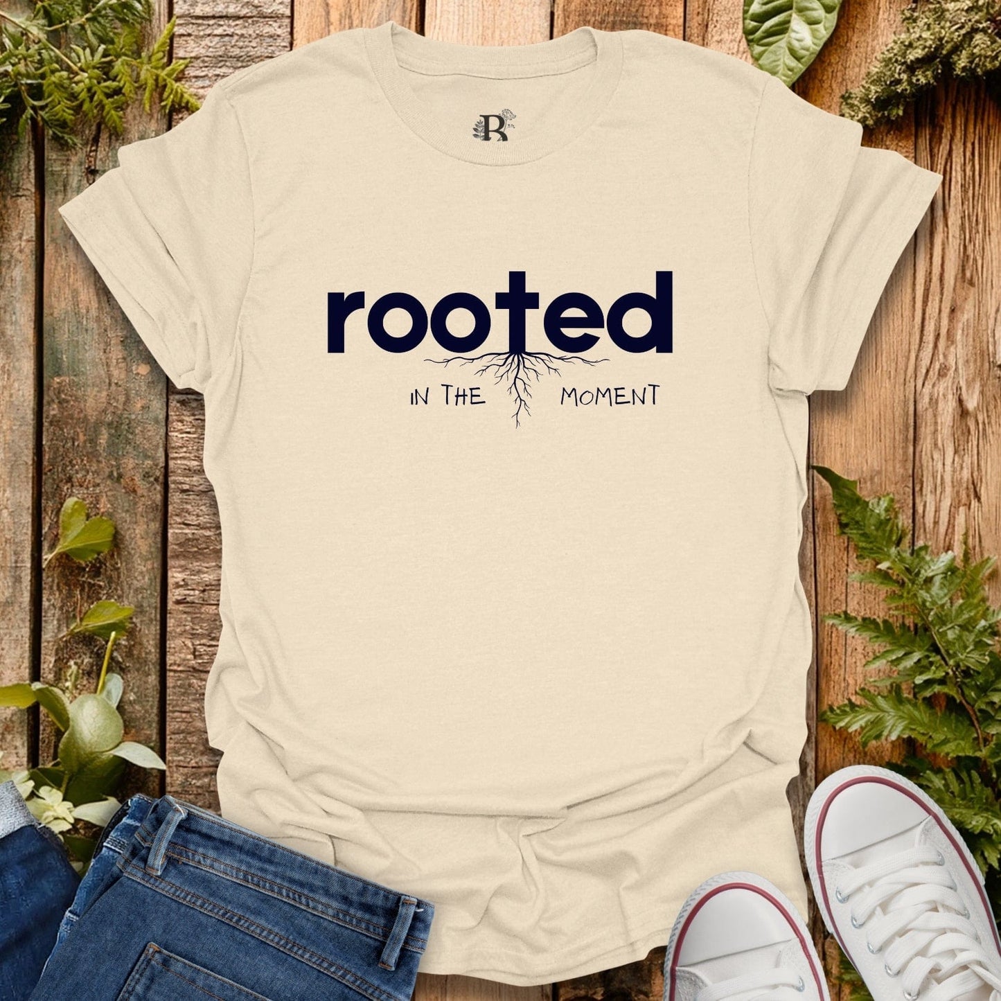 Beige t-shirt with 'rooted in the moment' text on a wooden background with plants.