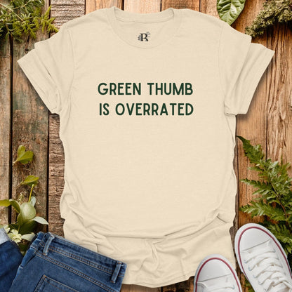Beige t-shirt with 'Green Thumb is Overrated' text on a wooden background with plants.