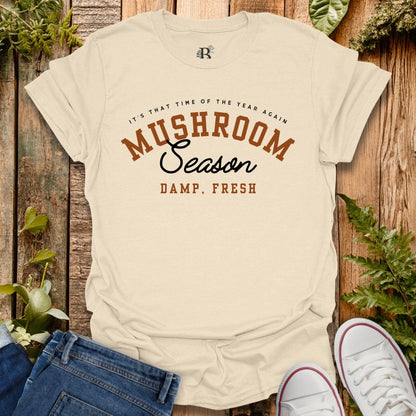 Beige t-shirt with 'Mushroom Season' text on a wooden background with plants.