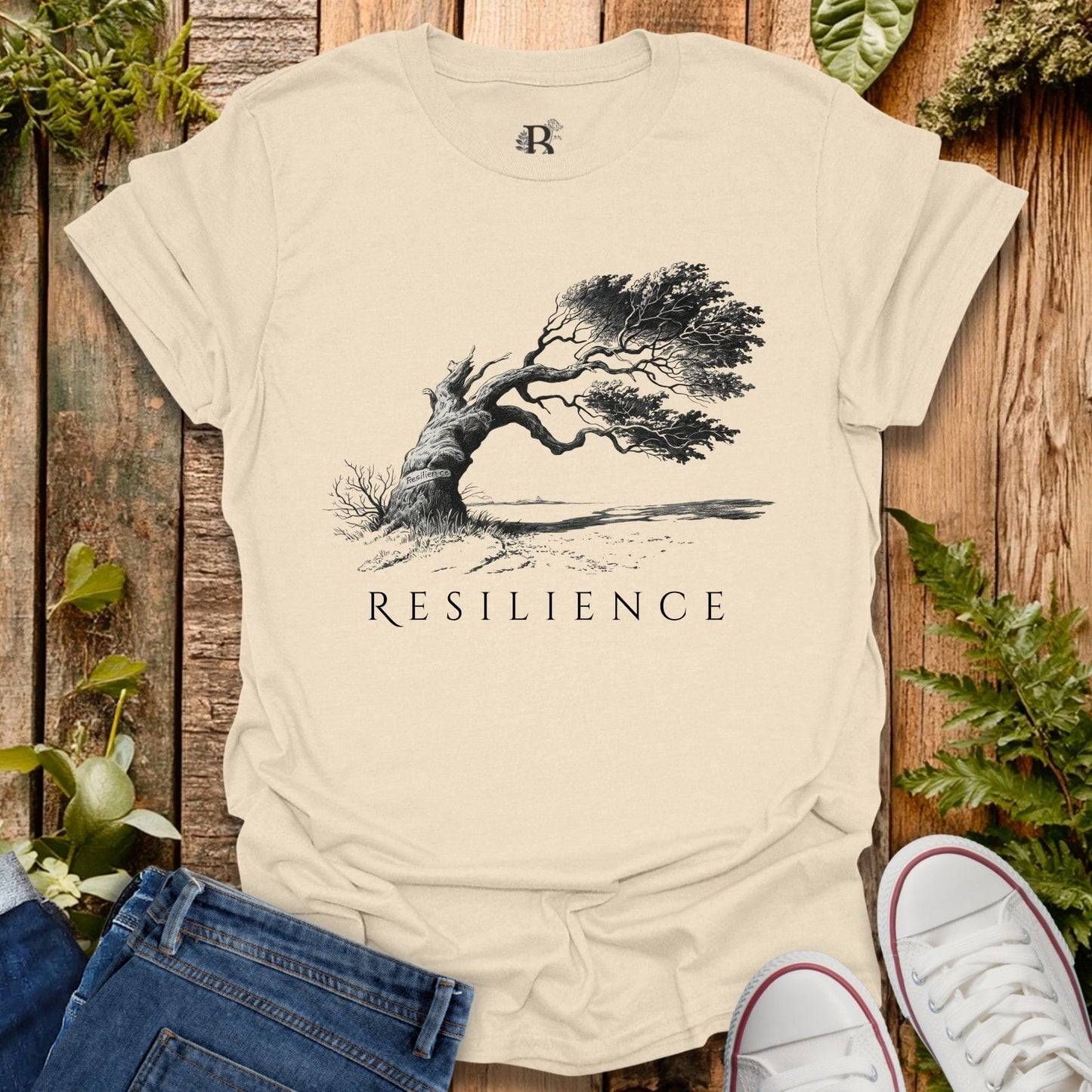 Beige t-shirt with 'Resilience' graphic and tree design on a wooden surface with plants.