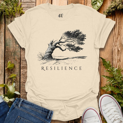 Beige t-shirt with 'Resilience' graphic and tree design on a wooden surface with plants.