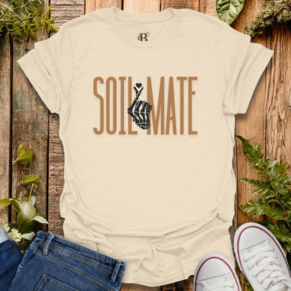 Beige t-shirt with 'SOIL MATE' text and hand symbol on a wooden background with plants.