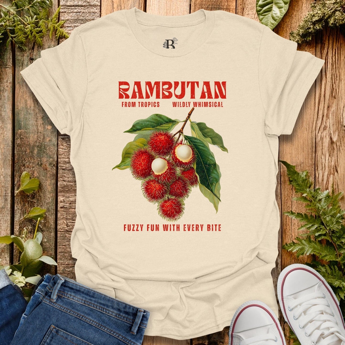 Beige t-shirt with rambutan graphic and text on a wooden surface with plants.