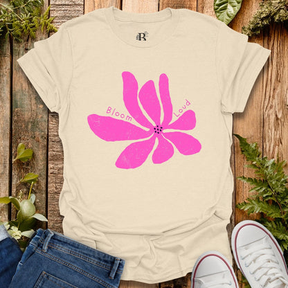 Beige t-shirt with a pink flower graphic and 'Bloom Loud' text on a wooden surface with plants.