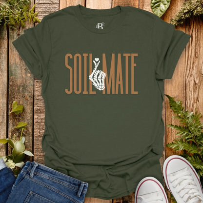 Green t-shirt with 'SOIL MATE' design on a wooden surface with plants.