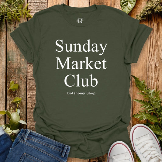 Green t-shirt with 'Sunday Market Club' text on a wooden surface with plants.