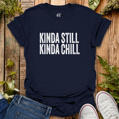 Navy blue t-shirt with 'KINDA STILL KINDA CHILL' text on a wooden surface with jeans and sneakers.