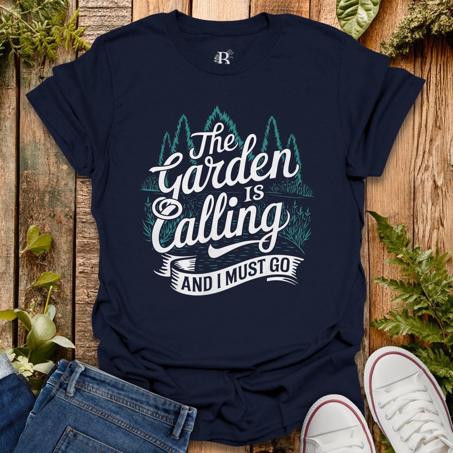 Navy blue t-shirt with 'The Garden is Calling and I Must Go' text on a wooden surface with plants.