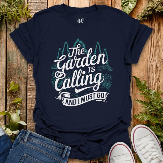 Navy blue t-shirt with 'The Garden is Calling and I Must Go' text on a wooden surface with plants.