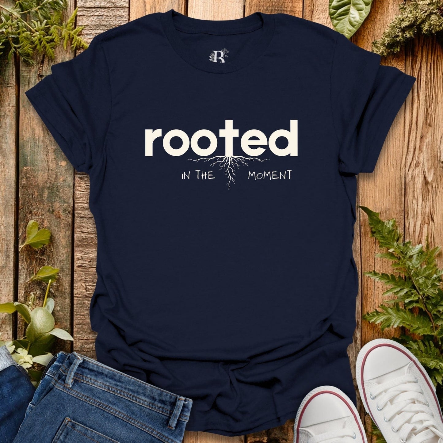 Navy blue t-shirt with 'rooted in the moment' text on a wooden surface with plants.