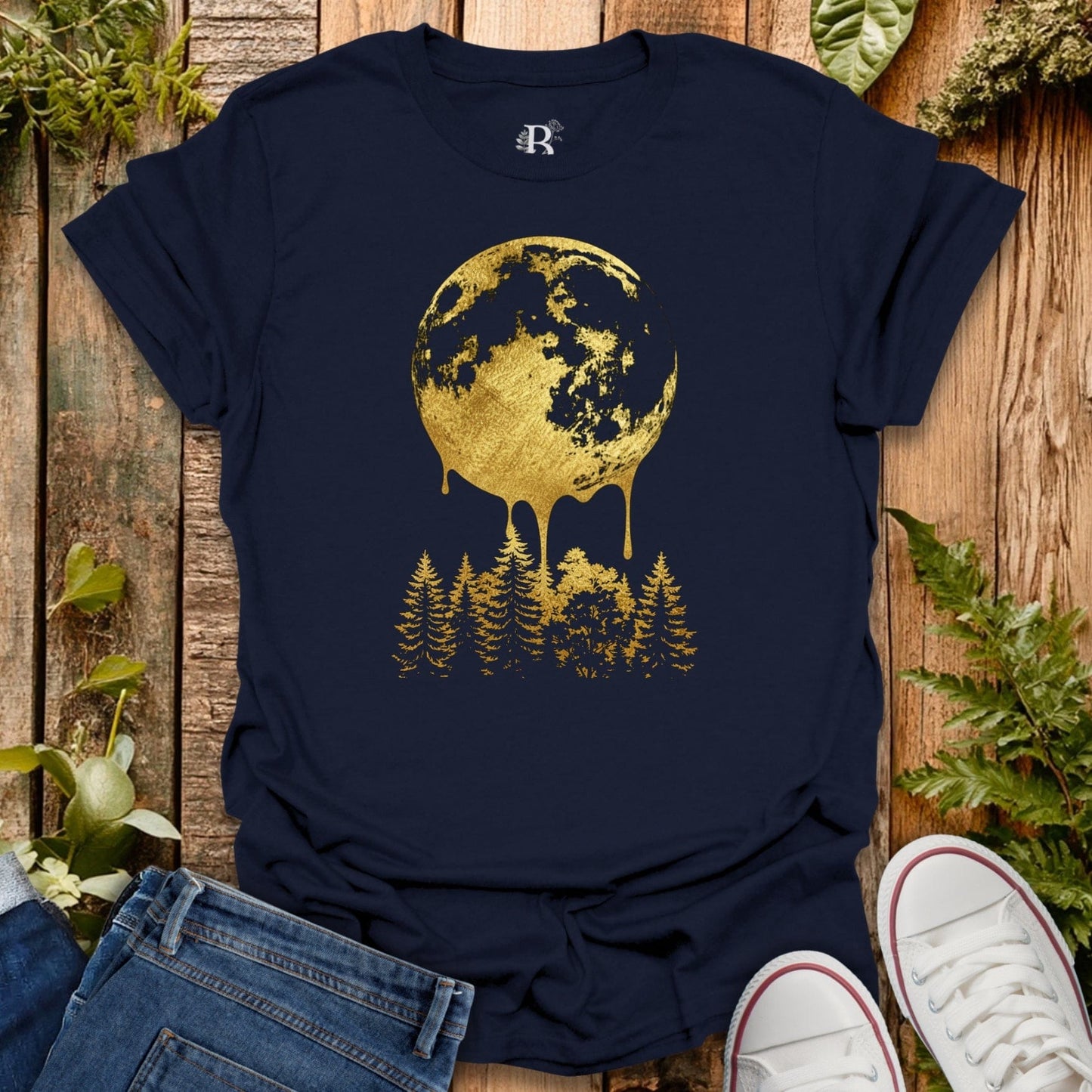 Navy blue t-shirt with gold moon and tree design on a wooden surface with plants.