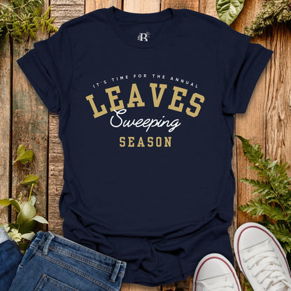 Navy blue t-shirt with 'Leaves Sweeping Season' text on a wooden surface with leaves and shoes.
