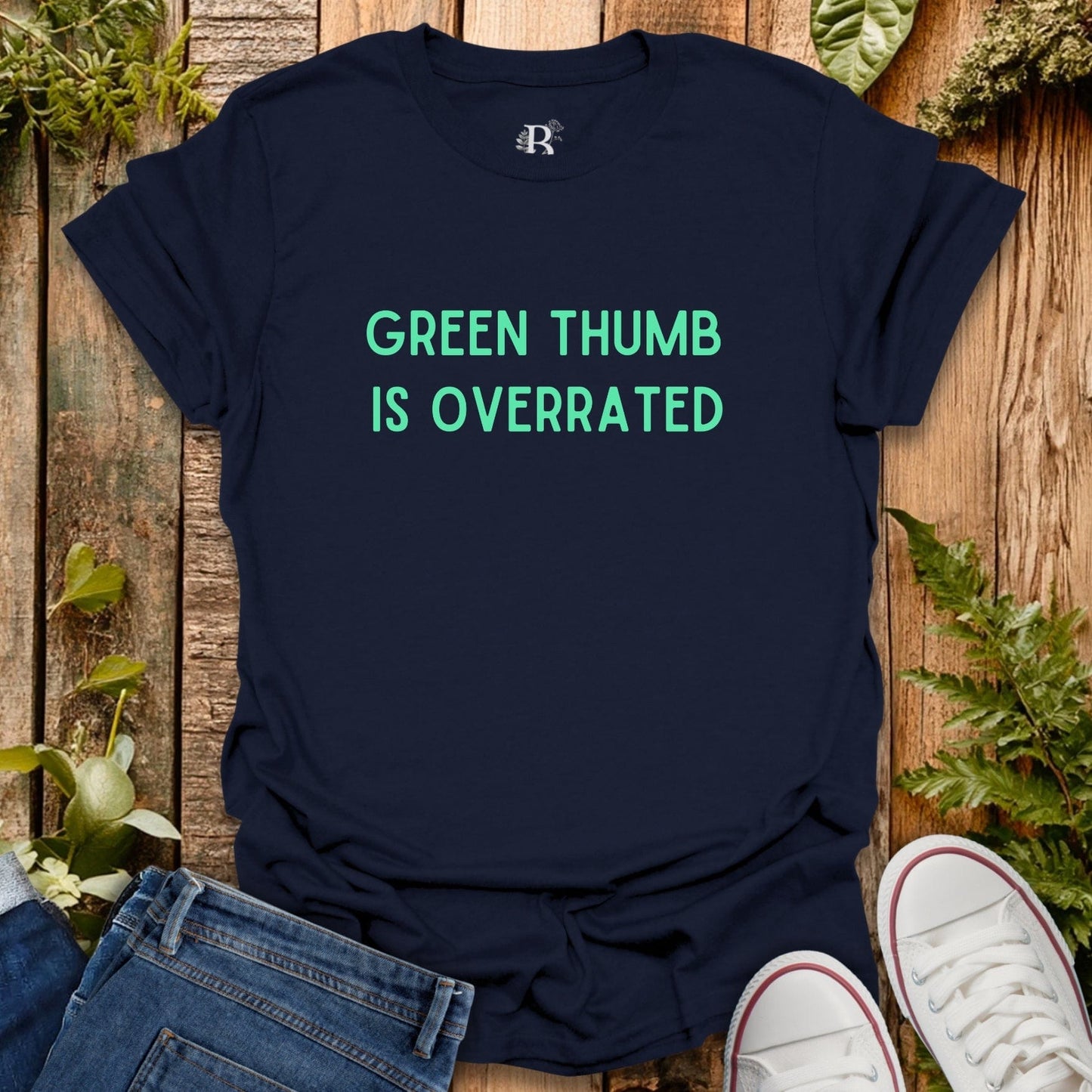 Navy blue t-shirt with 'Green Thumb is Overrated' text on a wooden surface with plants.
