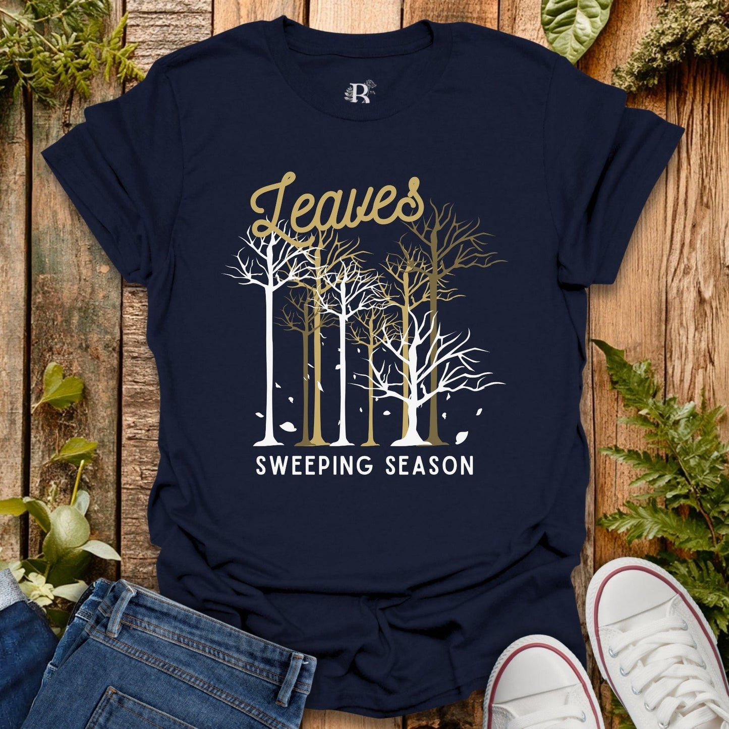 Navy blue t-shirt with 'Leaves Sweeping Season' design on a wooden surface with plants.