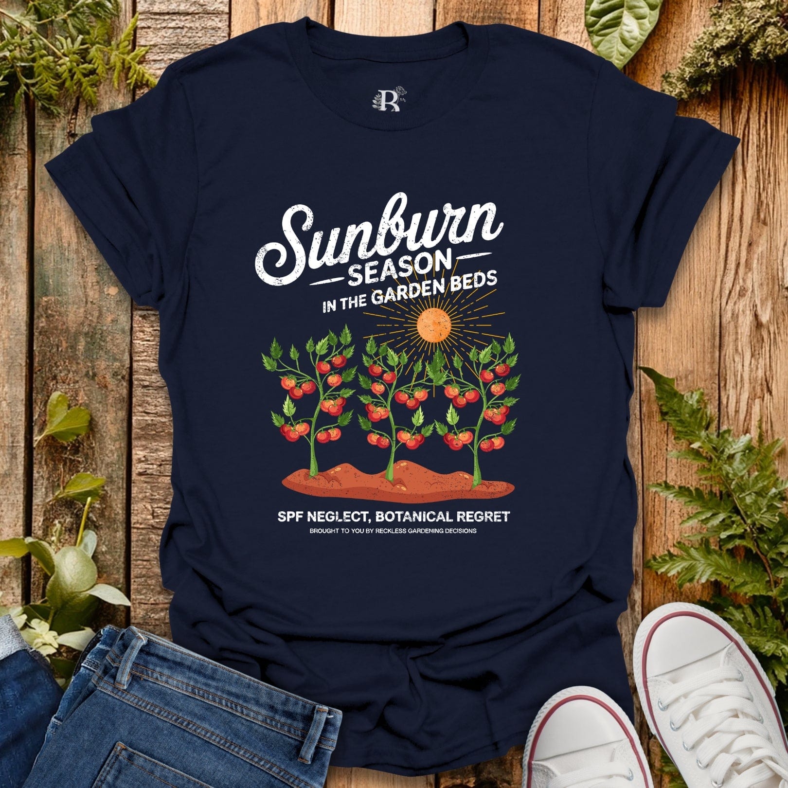 Navy blue t-shirt with 'Sunburn Season in the Garden Beds' graphic on a wooden surface with plants.