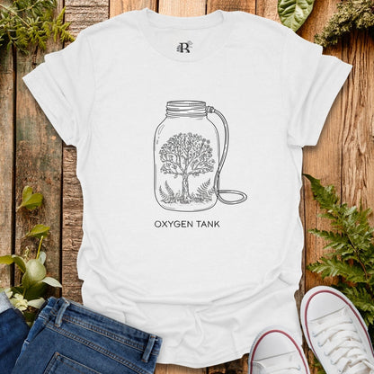White t-shirt with a tree design inside a jar labeled 'Oxygen Tank' on a wooden surface with plants.