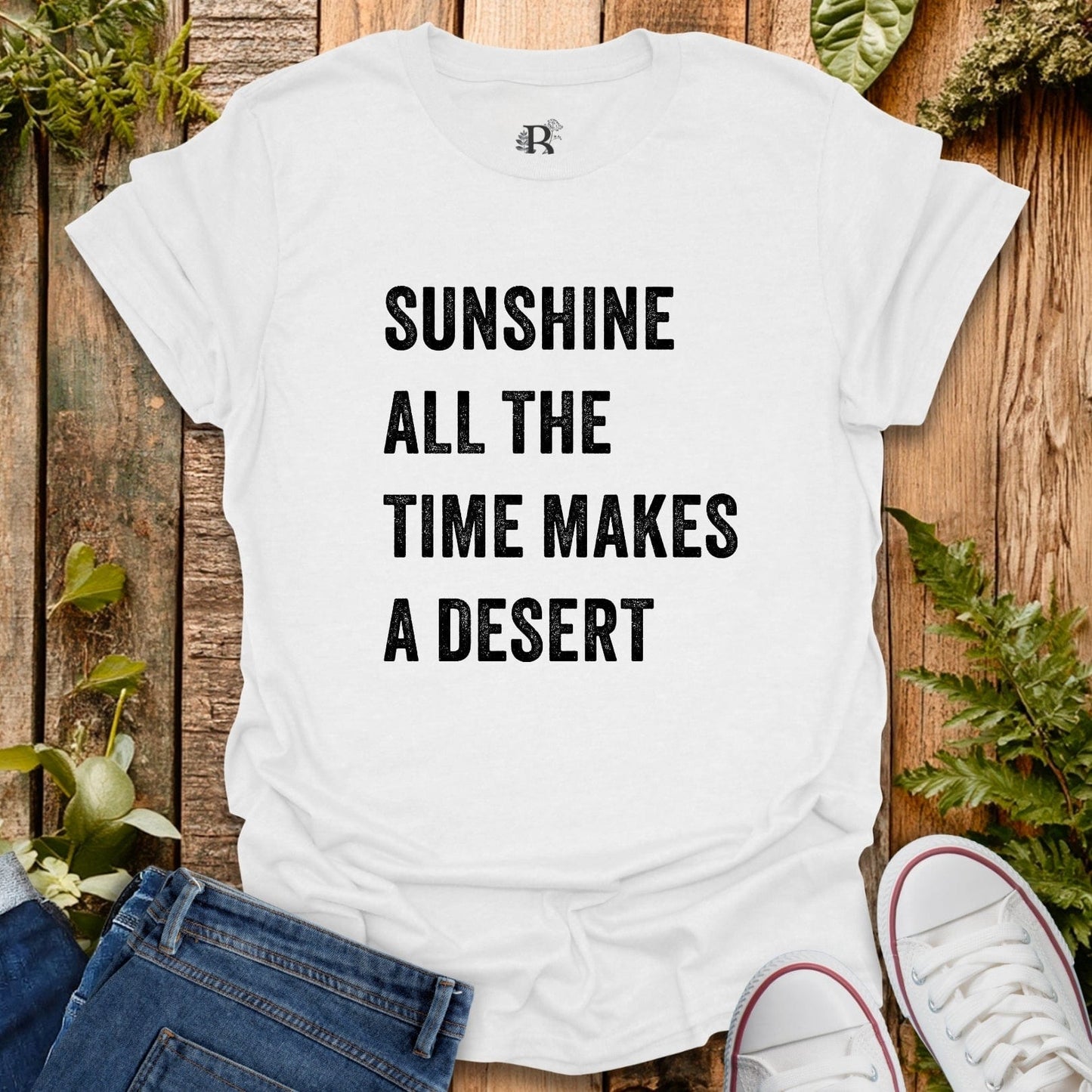 White t-shirt with text 'Sunshine all the time makes a desert' on a wooden surface with plants.