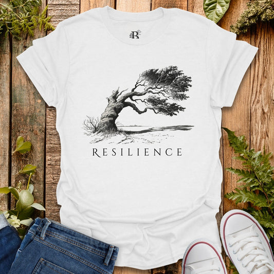 White t-shirt with a black tree graphic and 'RESILIENCE' text on a wooden surface with plants.