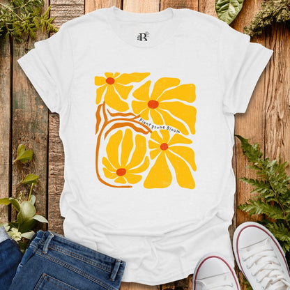 White t-shirt with yellow floral design on a wooden surface with plants.