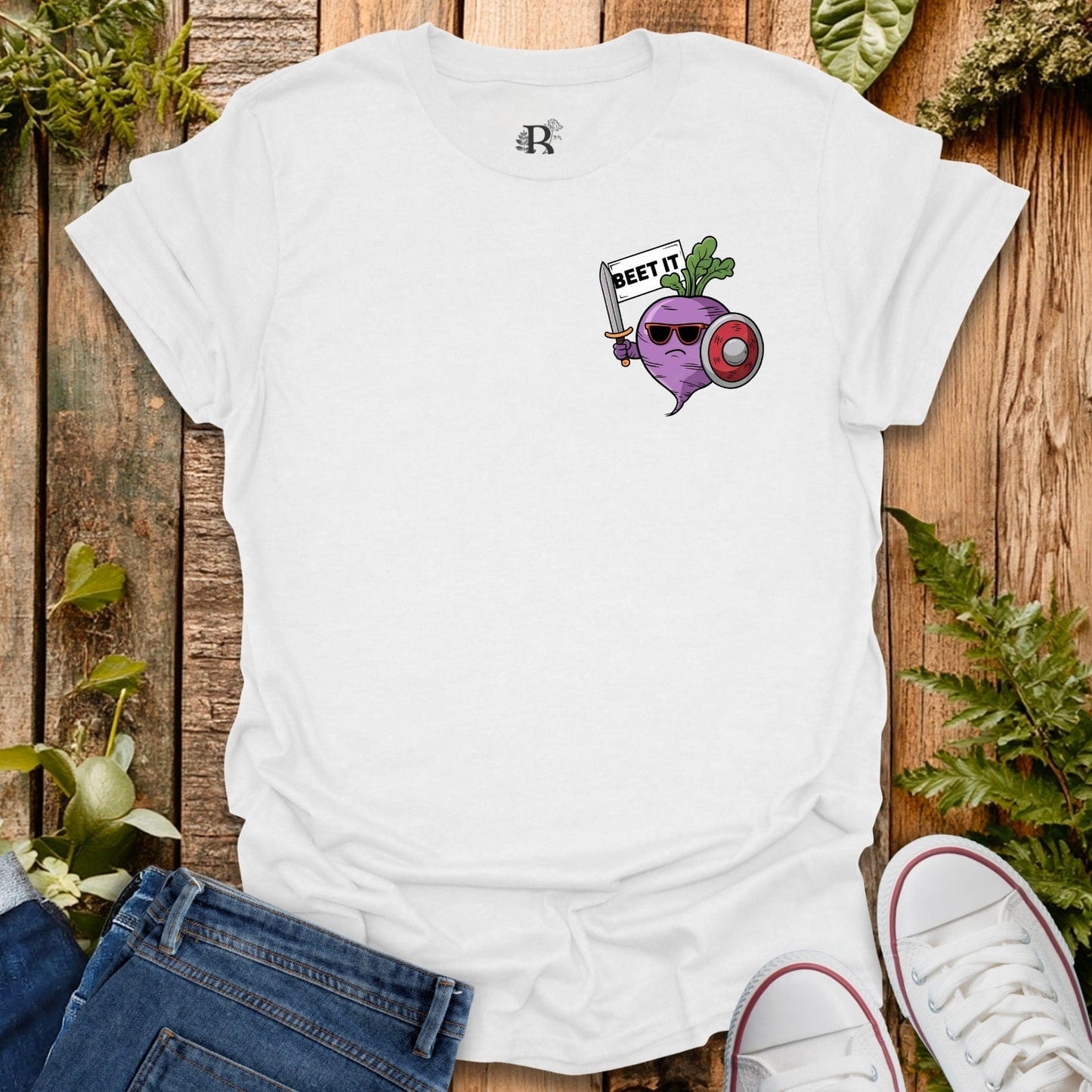 White t-shirt with a graphic design of a purple beet character holding a sword, placed on a wooden surface with plants.