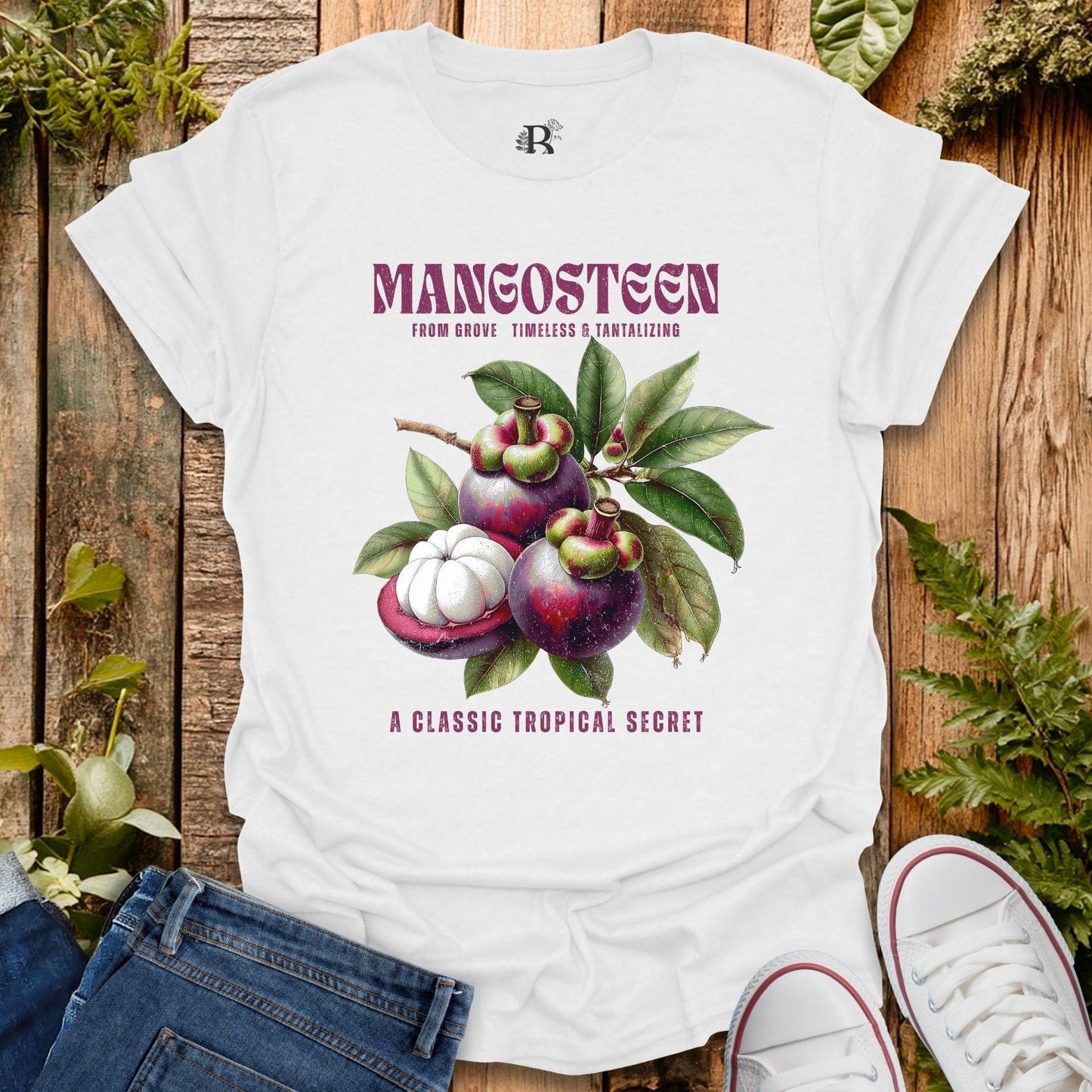 White t-shirt with mangosteen graphic and text on a wooden surface with plants.