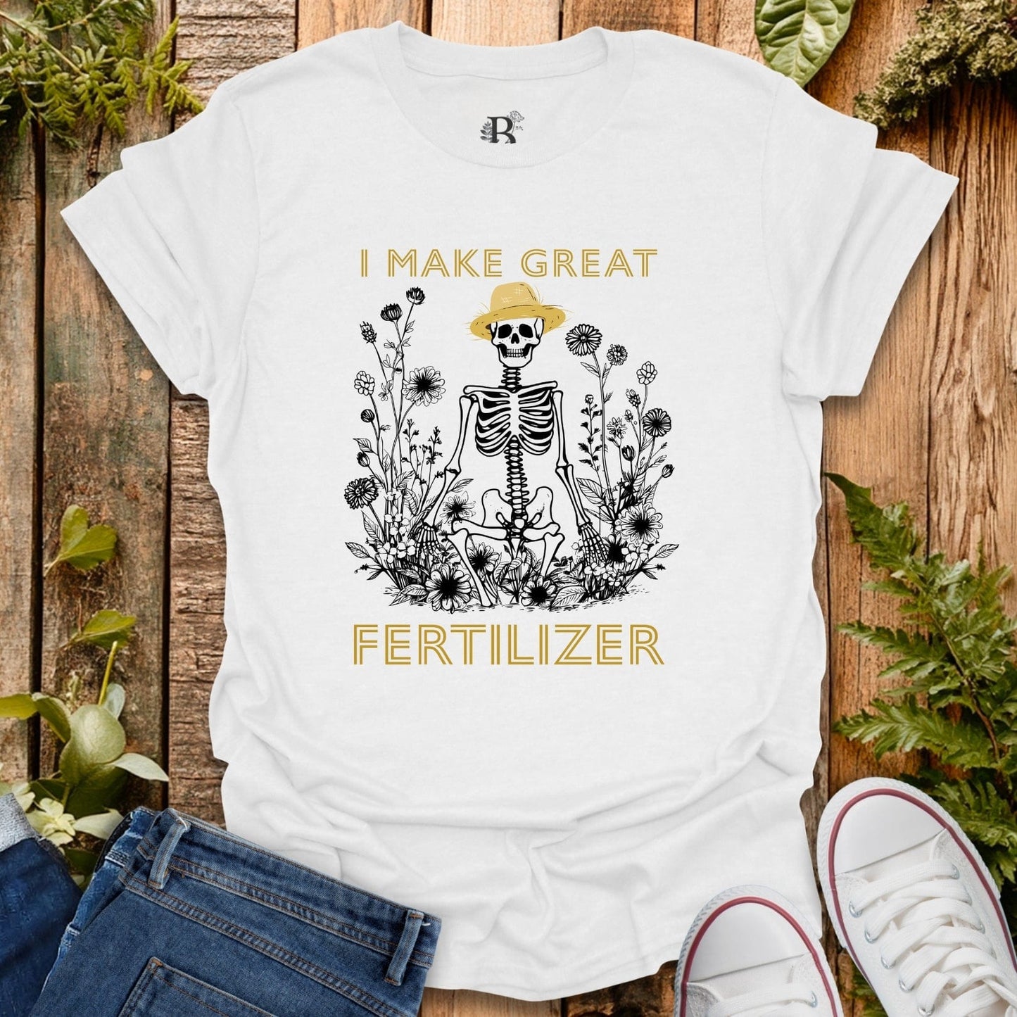 White t-shirt with skeleton graphic and text 'I MAKE GREAT FERTILIZER' on a wooden surface with plants.