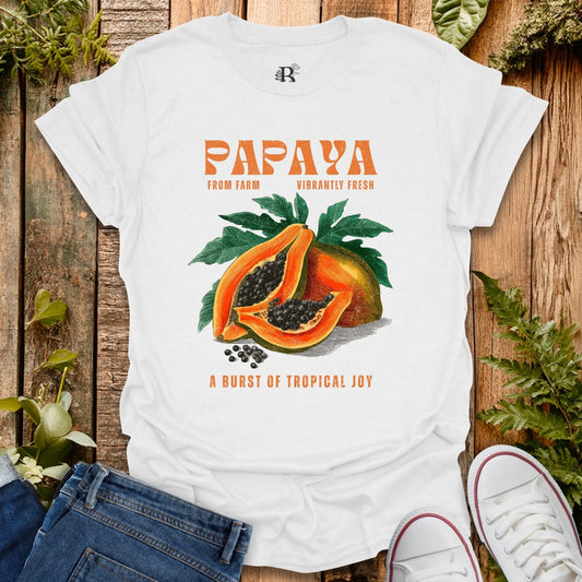 White t-shirt with papaya graphic and text on a wooden surface with plants.
