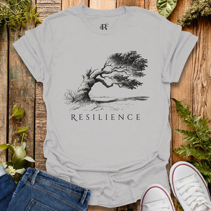 Gray t-shirt with 'Resilience' graphic and tree design on a wooden surface with plants.
