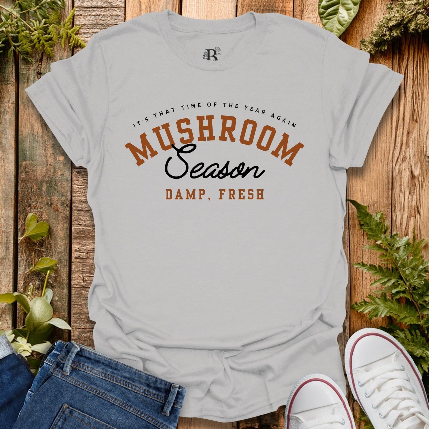 Gray t-shirt with 'Mushroom Season' text on a wooden background with plants.